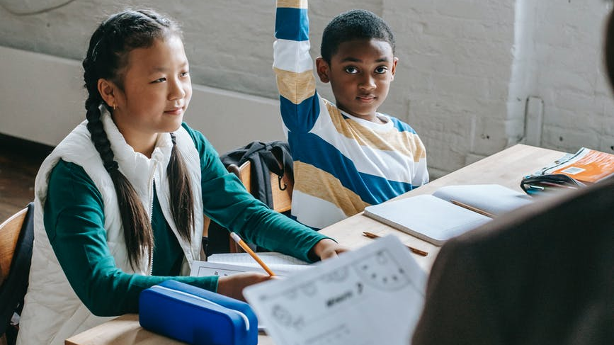 Photo of two children in a classroom. One has their hand up