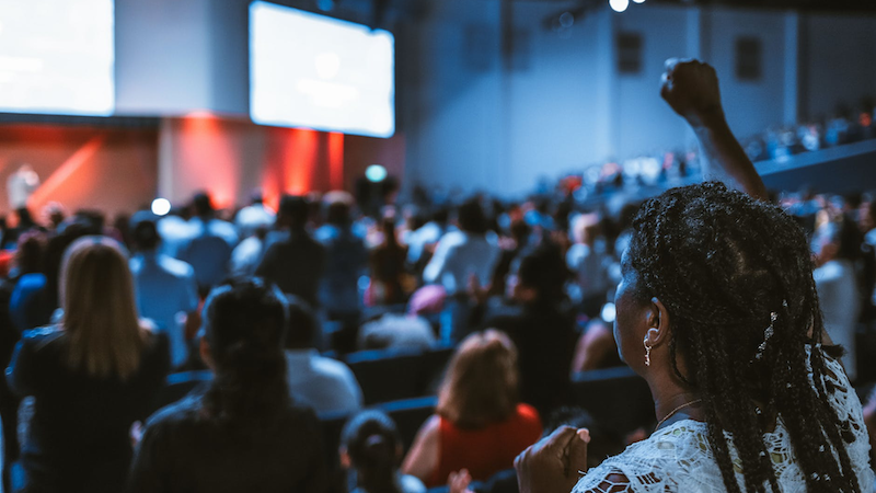 A view of a stage from the back of the room. A Black woman is prominently visible in the foreground, watching the presentation