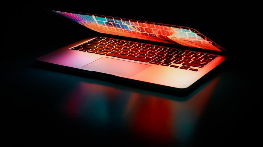 Photo of a laptop in a dark room, half open so the keyboard is illuminated in bright colours by the screen